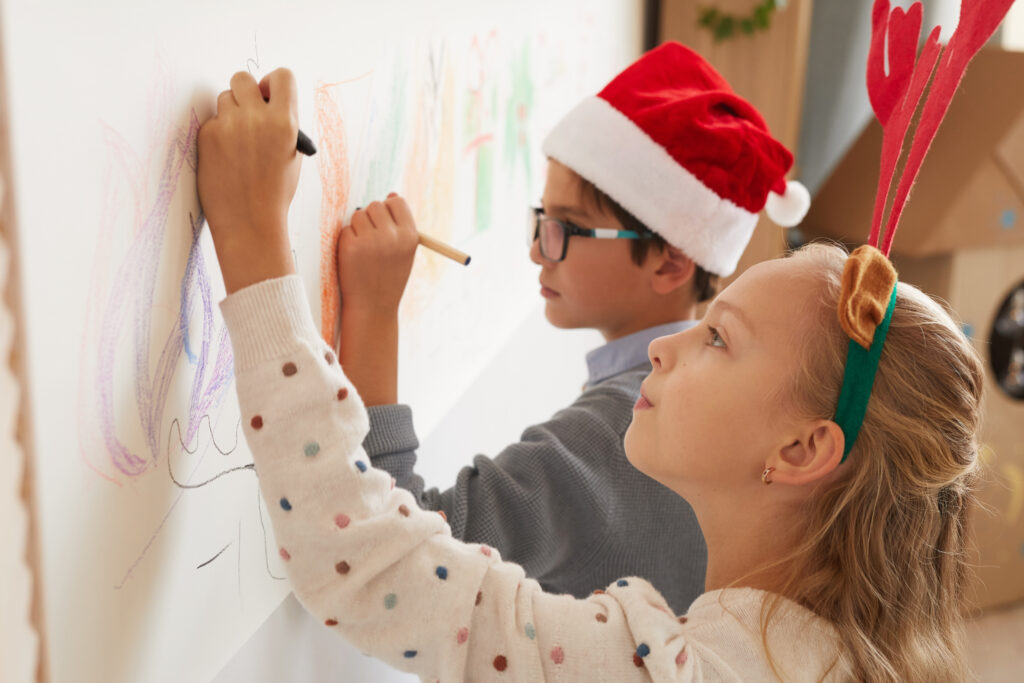 Side view portrait of boy and girl drawing on walls while wearing Santa hats and antlers for Christmas, copy space